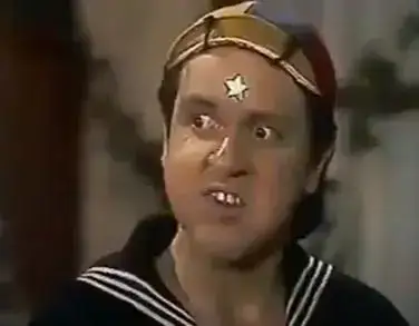 Personagem Quico do seriado Chaves com expressão de raiva, usando seu tradicional gorro colorido e uniforme de marinheiro azul com listras brancas, e com uma estrela branca colada na testa, em cena de fundo desfocado.