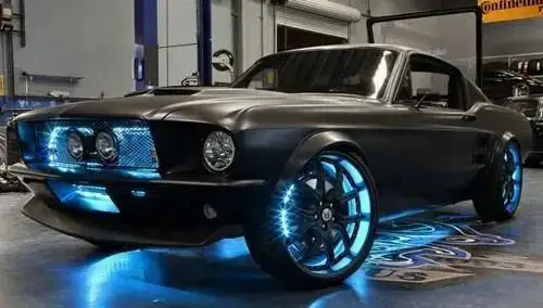 Um carro esportivo clássico, estilo muscle car, com pintura preta fosca e visual altamente personalizado. Está em uma garagem iluminada, com luzes de LED azuis brilhando sob a carroceria, nas rodas e na grade frontal. As rodas são grandes, com aros cromados e iluminação azul nos raios. O visual transmite potência, estilo futurista e destaque visual extremo.