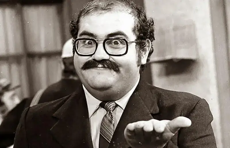 Senhor Barriga provando que Wishful Thinking não funciona. "Homem de bigode e óculos grandes, vestindo terno e gravata, com expressão facial caricata e um gesto de mão indicando convite ou questionamento, em uma fotografia em preto e branco.